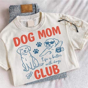 Dog Mom Club design displayed on a grey shirt outdoors with fun dog graphics and paw prints. Ideal for dog moms, animal lovers, and casual wear. Perfect for sublimation and print-on-demand designs.