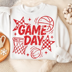 This ALT text describes a white sweatshirt lying on a plush cream blanket, showcasing a vivid red “GAME DAY” basketball graphic across the chest. The design includes distressed lettering, a hoop, bouncing balls, and checkered stars, all in matching red ink that pops against the white fabric. The sweatshirt is styled next to a wooden tray with winter décor, emphasizing a cozy sports-fan aesthetic. The large central print makes this piece perfect for basketball moms and spirited game-day wear.