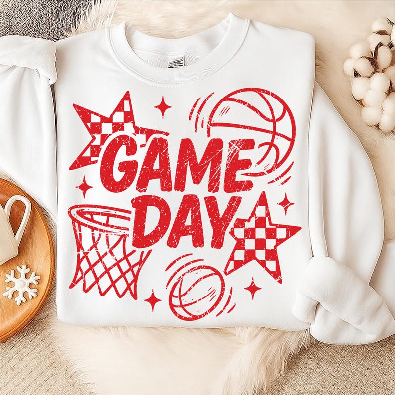 This ALT text describes a white sweatshirt lying on a plush cream blanket, showcasing a vivid red “GAME DAY” basketball graphic across the chest. The design includes distressed lettering, a hoop, bouncing balls, and checkered stars, all in matching red ink that pops against the white fabric. The sweatshirt is styled next to a wooden tray with winter décor, emphasizing a cozy sports-fan aesthetic. The large central print makes this piece perfect for basketball moms and spirited game-day wear.