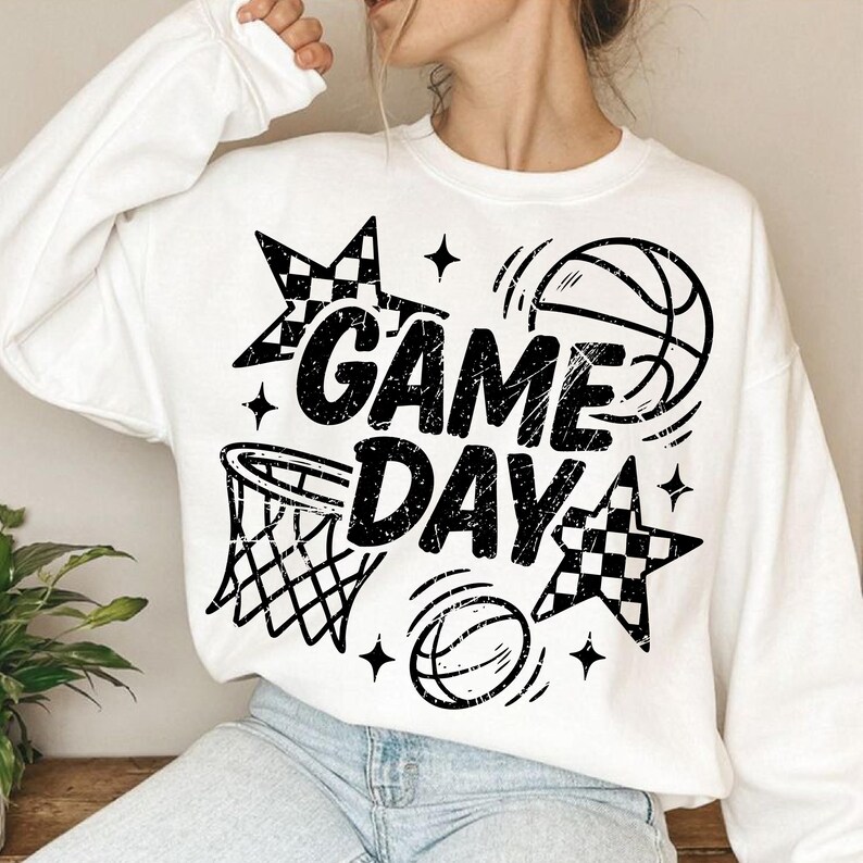 This ALT text describes a cozy white oversized sweatshirt worn by a woman, featuring a bold black “GAME DAY” basketball graphic centered on the chest. The design includes distressed black lettering, a basketball hoop illustration, bouncing basketballs, checkered star shapes, and small sparkles, giving it a sporty and energetic look. The large graphic fills most of the front of the sweatshirt, making it perfect for basketball moms, players, and fans dressing for game day.