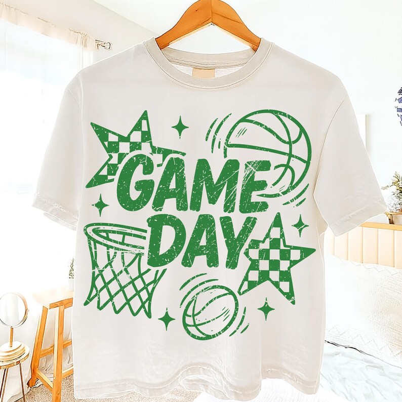 This ALT text describes a white unisex short-sleeve T-shirt hanging in a softly lit bedroom, featuring a large green “GAME DAY” basketball print. The graphic includes distressed lettering, a hoop, motion-style basketball sketches, checkered star shapes, and sparkles. The centered design fills most of the shirt’s front, showing ideal scale for everyday wear or team-spirit outfits. The green colorway works well for school teams or fans wanting a bold, sporty, game-day look.