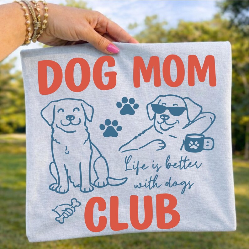 Dog Mom Club design displayed on a grey shirt outdoors with fun dog graphics and paw prints. Ideal for dog moms, animal lovers, and casual wear. Perfect for sublimation and print-on-demand designs.