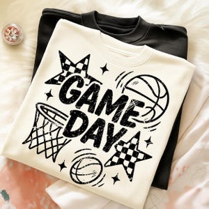 This ALT text describes a cream-colored sweatshirt folded neatly to reveal a black “GAME DAY” basketball design on the front. The graphic includes distressed lettering, a hoop illustration, basketball outlines, checkered stars, and sparkle accents. The sweatshirt is placed on a soft faux-fur surface with a black sweatshirt layered underneath, showcasing color contrast and fabric texture. The design spans most of the chest area, making it ideal for basketball moms, team supporters, and sport