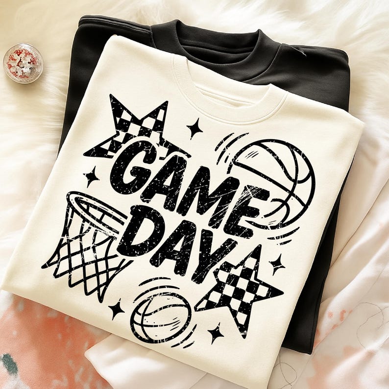 This ALT text describes a cream-colored sweatshirt folded neatly to reveal a black “GAME DAY” basketball design on the front. The graphic includes distressed lettering, a hoop illustration, basketball outlines, checkered stars, and sparkle accents. The sweatshirt is placed on a soft faux-fur surface with a black sweatshirt layered underneath, showcasing color contrast and fabric texture. The design spans most of the chest area, making it ideal for basketball moms, team supporters, and sport