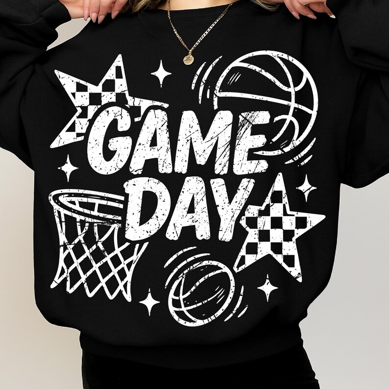 This ALT text describes a black oversized sweatshirt featuring a striking white “GAME DAY” graphic printed across the front. The distressed white letters are accented with white basketball outlines, a basketball net illustration, checkered stars, and sparkles, creating a high-contrast sporty design. The model is holding the neckline, showing how the large design spans from the upper chest to the lower midsection.