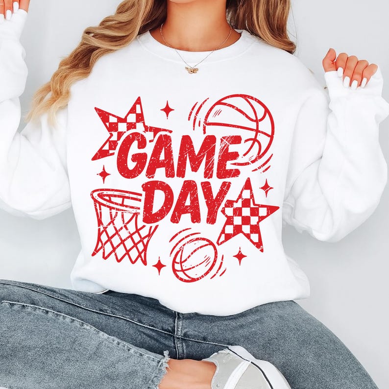 This ALT text describes a bright white sweatshirt styled with a large red “GAME DAY” basketball print covering the chest area. The distressed red lettering is paired with a basketball hoop, bouncing basketballs, checkered stars, and sparkles, all in matching red linework. The woman wearing the sweatshirt is seated comfortably in jeans, highlighting the width and full coverage of the design. Ideal for basketball fans, team moms, and sporty outfits