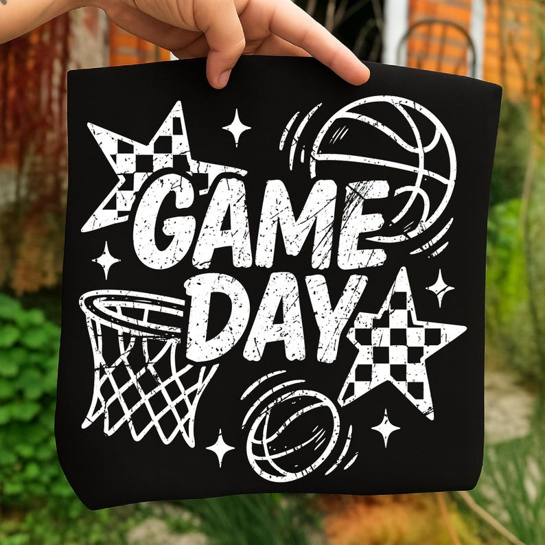This ALT text describes a black sheet held outdoors by a hand, printed with a white “GAME DAY” basketball graphic intended for heat transfer crafting. The design features distressed block letters, a basketball hoop, bouncing basketballs, checkered stars, and sparkle details. The outdoor background contrasts with the sheet, highlighting the clarity and scale of the graphic. The transfer is sized to cover the front of a sweatshirt or T-shirt, ideal for DIY basketball apparel, team gifts