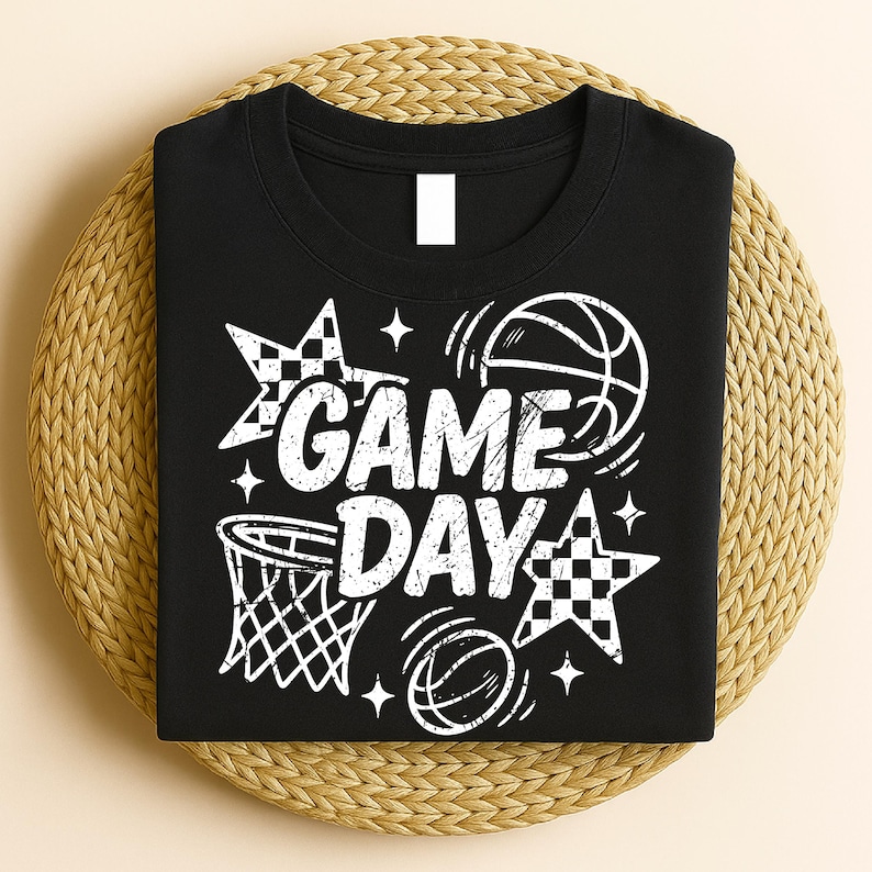This ALT text describes a neatly folded black unisex T-shirt displayed on a woven round mat, showcasing the white “GAME DAY” basketball design on the front. The print features distressed block letters, outlines of basketballs in motion, a basketball hoop, checkered stars, and sparkle accents. The arrangement emphasizes the full size and placement of the graphic when worn. The crisp white ink contrasts sharply with the black shirt, making the sporty design perfect for basketball lovers.