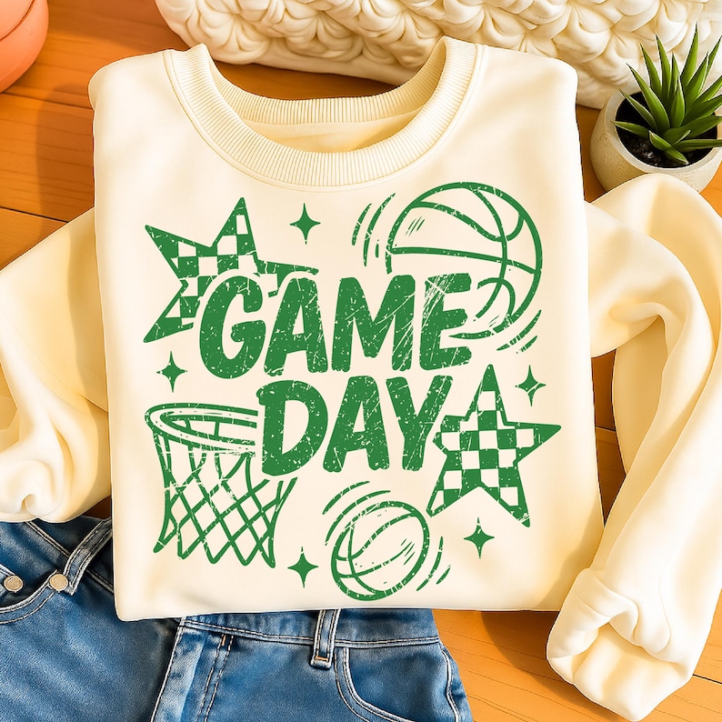 This ALT text describes a soft cream sweatshirt displayed on a wooden surface next to denim jeans, featuring a bold green “GAME DAY” design. The graphic includes distressed lettering, sketched basketball icons, a hoop, star shapes with checkered patterns, and small sparkles. The print covers a wide central area of the sweatshirt, showing how it fits on the chest when worn. This sporty basketball design is perfect for team moms, school spirit wear, and game-day outfits in team-matching green