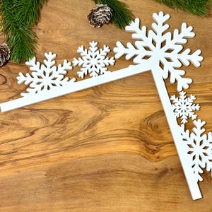 May include: White snowflake-shaped decorations arranged in a corner design. The snowflakes are of varying sizes and are placed on a wooden surface. Green pine needles and pine cones are in the background.