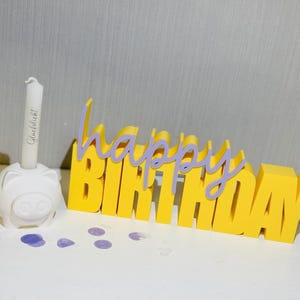 Puede incluir: Un letrero amarillo de "Happy Birthday" con escritura púrpura superpuesta. Un portavelas blanco con forma de cerdo sostiene una vela blanca con escritura. Confeti púrpura salpica la superficie.