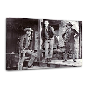 Peut inclure: Impression sur toile en noir et blanc représentant trois hommes en tenue de western. Les hommes se tiennent sur un porche en bois devant un bâtiment en briques. Ils portent des chapeaux de cow-boy, des gilets et des jeans.