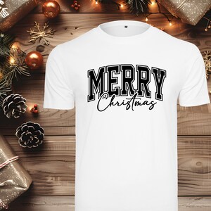 Könnte beinhalten: Weißes T-Shirt mit dem Text "MERRY Christmas" in schwarzer Schrift im College-Stil. Das Wort "MERRY" ist fett und in Blockbuchstaben gehalten, während "Christmas" kursiv geschrieben ist. Das Shirt liegt auf einer Holzoberfläche mit Weihnachtsdekorationen.