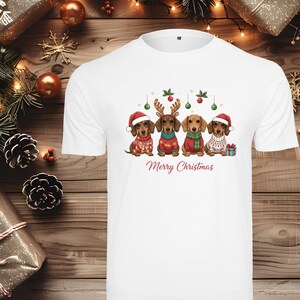 Könnte beinhalten: Weißes T-Shirt mit einem weihnachtlichen Design. Es zeigt vier Dackel in festlichen Outfits, darunter Weihnachtsmützen und Rentiergeweihe. Die Worte "Merry Christmas" sind unter den Hunden aufgedruckt.
