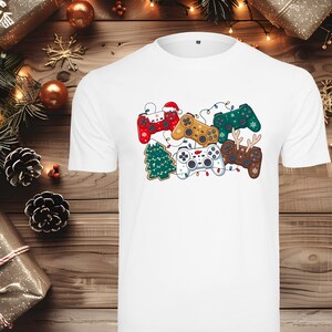 Könnte beinhalten: Weißes T-Shirt mit einem festlichen Design von dekorierten Gamecontrollern. Die Controller sind in verschiedenen Farben, darunter Rot, Gold und Grün, mit weihnachtlichen Dekorationen. Ein Weihnachtsbaum ist ebenfalls enthalten.