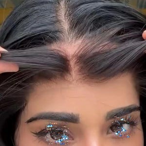 Può includere: Primo piano del viso di una persona con capelli scuri divisi a metà. La persona tiene indietro i capelli, rivelando il cuoio capelluto. Gli occhi sono adornati di glitter e le sopracciglia sono ben definite.