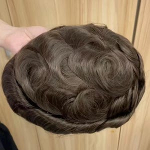 Op de afbeelding: Een bruin haarstuk met een golvende textuur. Het haarstuk heeft een donkerbruine kleur en lijkt gemaakt te zijn van synthetisch of menselijk haar. Het haar is gestyled in losse golven.