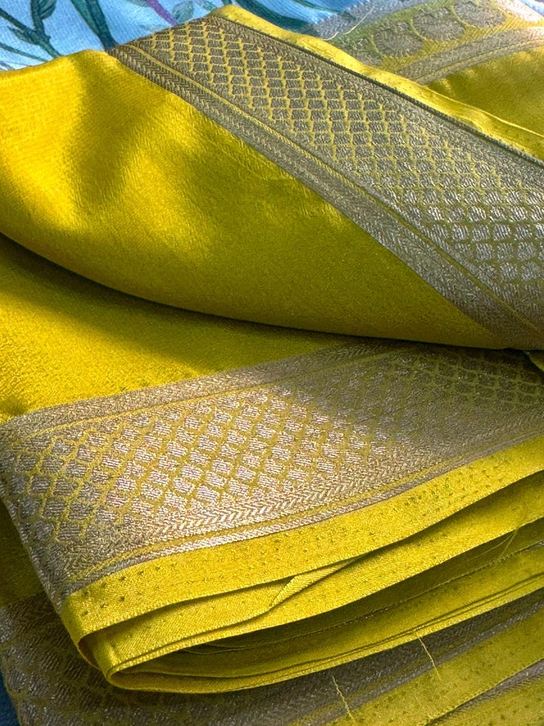 Op de afbeelding: Close-up van een gevouwen, heldergele stof met een glinsterende textuur. De stof heeft een decoratieve rand met een geometrisch patroon in een contrasterende zilvergrijze kleur. De plooien cre&euml;ren lagen.