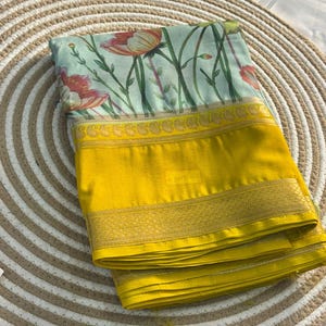 Op de afbeelding: Een gevouwen textiel met een bloemenpatroon van rode en oranje bloemen op een lichtblauwe achtergrond. De rand is heldergeel met een gouden decoratieve rand. De stof is netjes gevouwen.