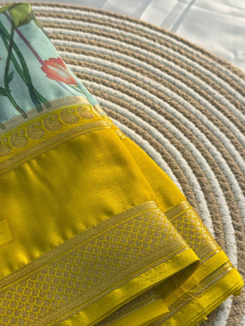 Op de afbeelding: Een gevouwen gele textiel met een decoratieve gouden rand. Het textiel heeft een bloemenpatroon in tinten blauw, groen en roze. De stof is gedrapeerd over een geweven, cirkelvormig oppervlak.