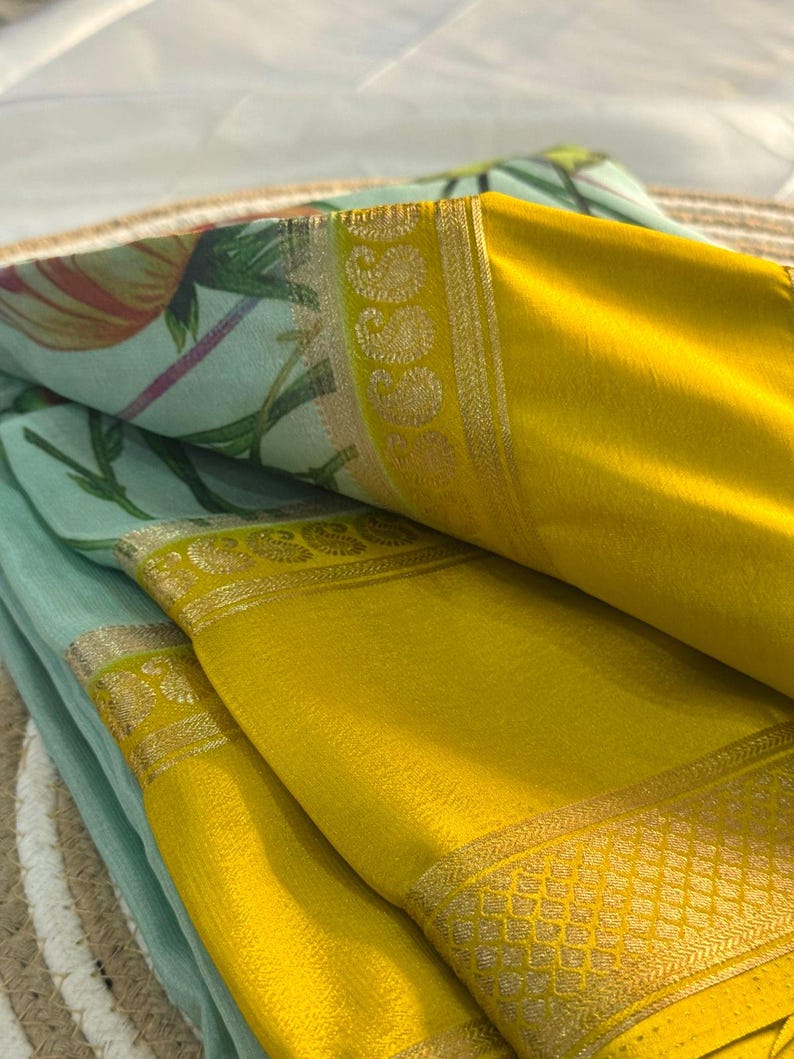 Op de afbeelding: Close-up van gevouwen stof met een bloemenprint in groen, roze en geel. De stof heeft een felgele rand met gouden details. De lagen stof zijn zo gerangschikt dat de contrasterende kleuren en texturen worden getoond.