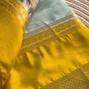 Op de afbeelding: Een gevouwen textiel met een heldergele rand en gouden details. De hoofdstof is lichtblauw met een bloemenpatroon in roze, oranje en groen. De stof heeft een zijdeachtige glans, met een decoratieve rand.