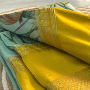Op de afbeelding: Close-up van gevouwen stof met een bloemenprint in groen, roze en geel. De stof heeft een felgele rand met gouden details. De lagen stof zijn zo gerangschikt dat de contrasterende kleuren en texturen worden getoond.
