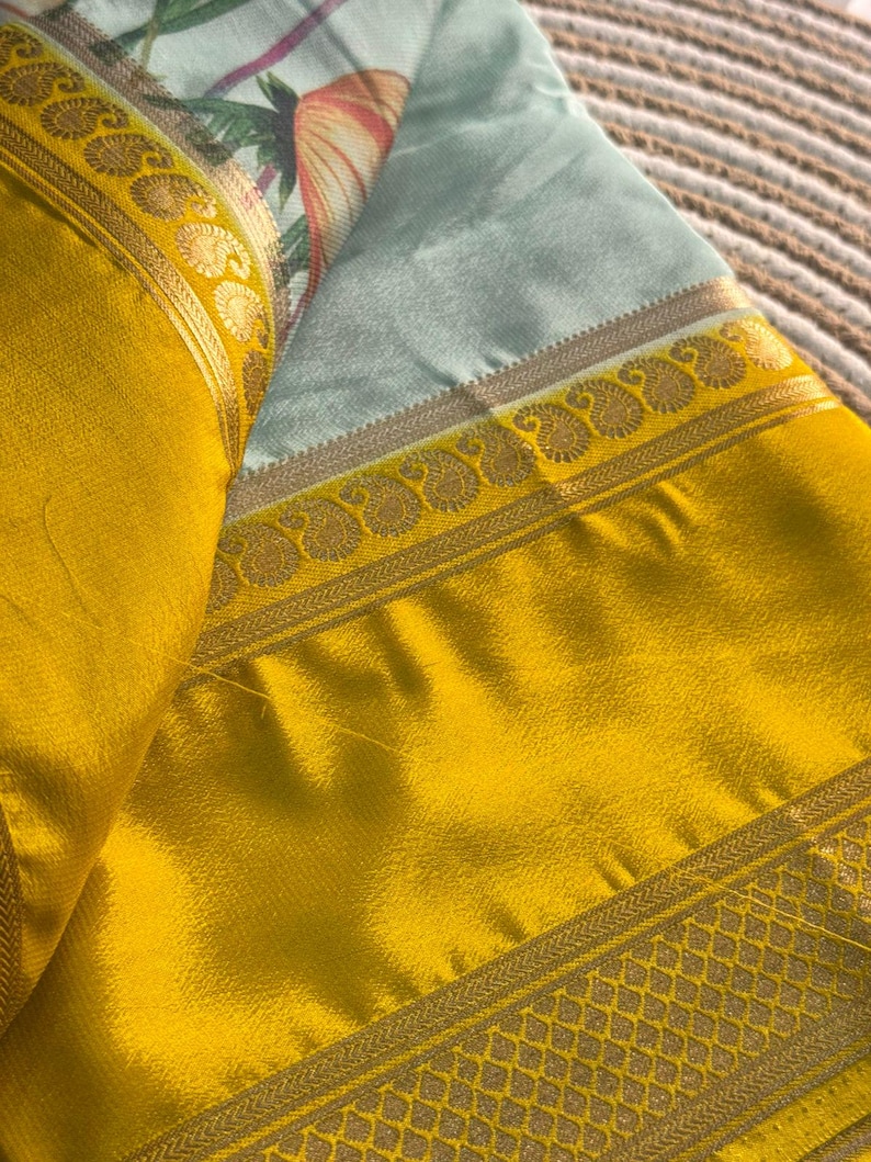 Op de afbeelding: Een gevouwen textiel met een heldergele rand en gouden details. De hoofdstof is lichtblauw met een bloemenpatroon in roze, oranje en groen. De stof heeft een zijdeachtige glans, met een decoratieve rand.