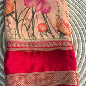 May include: A folded textile featuring a floral print in shades of peach, pink, and orange. The design includes a red border with gold accents. The fabric is folded, showcasing the pattern and color contrast.