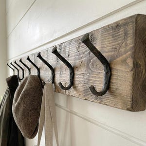 May include: A rustic wooden coat rack with eight black metal hooks. The wood has a natural, weathered appearance. A green jacket, a brown tweed cap, and a beige tote bag hang from the hooks, suggesting a functional entryway decor.