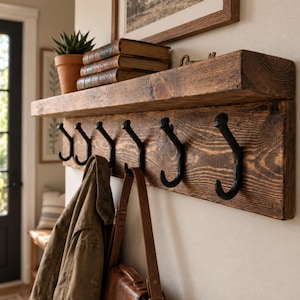 May include: A wall-mounted wooden shelf featuring a thick, dark-stained plank with visible wood grain. Attached to the front are six black metal hooks. A stack of three books and a small potted succulent sit on the top surface of the shelf.