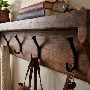 May include: A rustic wooden wall shelf featuring four dark metal hooks. The shelf holds three stacked books and a magnifying glass. A cream-colored knit fabric hangs from the leftmost hook, while a brown leather bag hangs from the center hook.