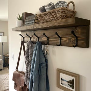 May include: A rustic, wooden wall shelf with a lower row of black metal hooks. The shelf holds a woven basket, rolled towels, books, and a potted plant. A leather bag, scarf, and denim jacket hang from the hooks.