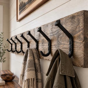 May include: A wall-mounted coat rack featuring a thick, rectangular piece of reclaimed brown wood with a visible grain. Seven black, forged metal hooks are attached to the wood, each secured with two screws. A plaid scarf and a jacket hang from hooks.