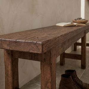 May include: A rectangular wooden bench with a dark brown, weathered finish and visible wood grain texture. The bench features four sturdy legs and a thick, solid top surface, showcasing a natural, aged appearance with distinct cracks and knots.