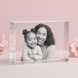 May include: A clear, rectangular crystal block displaying a black and white photograph of a smiling woman and a young child. The photo is set against a soft pink background, with a blurred floral element on the side.