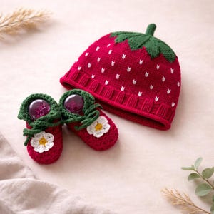 Peut inclure: Un bonnet et des chaussons pour bébé sur le thème de la fraise, tricotés en rouge. Le bonnet a une tige et des feuilles vertes, avec des détails de graines blanches. Les chaussons sont rouges avec des fleurs blanches et une bordure verte.