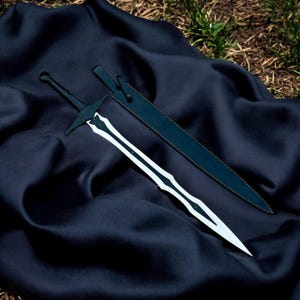 May include: A black and silver sword with a matching sheath rests on a dark blue fabric. The sword features a crossguard and a decorative design on the blade. The sheath is also black with a tan-colored stitch.