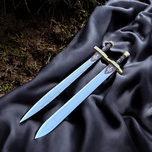 May include: Two fantasy swords with light blue blades and gold-colored hilts rest on a dark gray fabric. The hilts have a crossguard design and a brown grip. Each blade has a decorative pattern and a crest near the hilt.