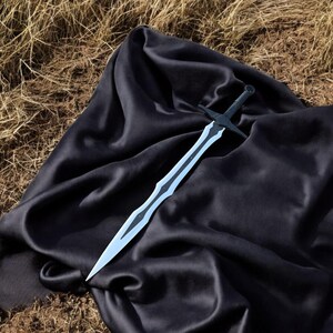 May include: A black and silver sword with a crossguard, resting on a black satin cloth. The blade has a light blue center and a decorative edge. The background is dry grass.