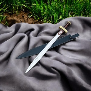 May include: A silver sword with a brown handle and gold accents, resting on a gray cloth. The sword has a decorative cross design on the blade and comes with a black sheath. The background features green grass.