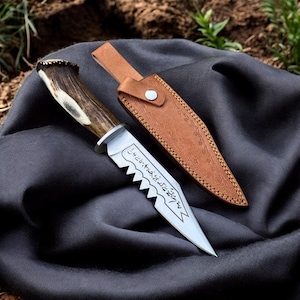 May include: A hunting knife with a serrated blade and a handle made of deer antler. The knife is accompanied by a brown leather sheath. The blade has etched text.