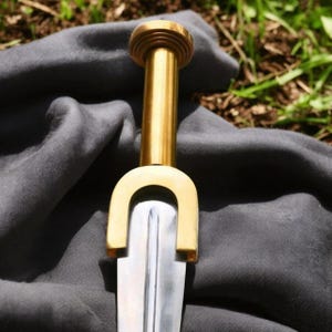 May include: Close-up of a sword with a polished silver blade and a gold-colored handle. The handle features a rounded pommel and a curved guard. The sword rests on a dark gray fabric, with a blurred green background.