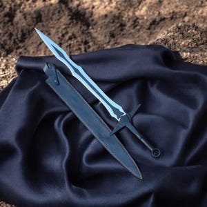 May include: A fantasy sword with a distinctive, light blue blade and a black hilt, resting on a dark blue cloth. The sword is paired with a black leather sheath, creating a striking contrast against the background.
