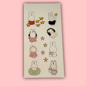 Könnte beinhalten: Ein Bogen mit Aufklebern mit Cartoon-Hasen in verschiedenen Posen. Einige Hasen tragen Kopfhörer, halten einen Sternenstab oder lesen ein Buch. Andere Aufkleber zeigen gelbe Entenküken und Sterne. Die Aufkleber befinden sich auf weißem Hintergrund.