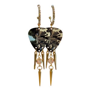 May include: A pair of gold-toned earrings. Each earring features a guitar pick-shaped pendant with a black and white image. The earrings have a dangling design with gold spikes and a clear bead. The top of the earrings have a curved design with small clear stones.