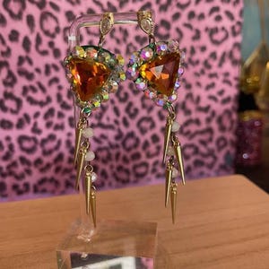 May include: A pair of ornate earrings on display. Each earring features a large, orange, heart-shaped gem surrounded by smaller, iridescent stones. Gold-colored spikes and beads dangle below. The earrings are displayed on a clear stand against a pink leopard print background.