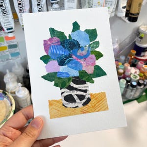 May include: A collage art piece featuring a bouquet of paper flowers in shades of blue, pink, and green. The flowers are arranged in a black and white striped vase with a yellow and brown patterned base. The artwork is set against a white background.