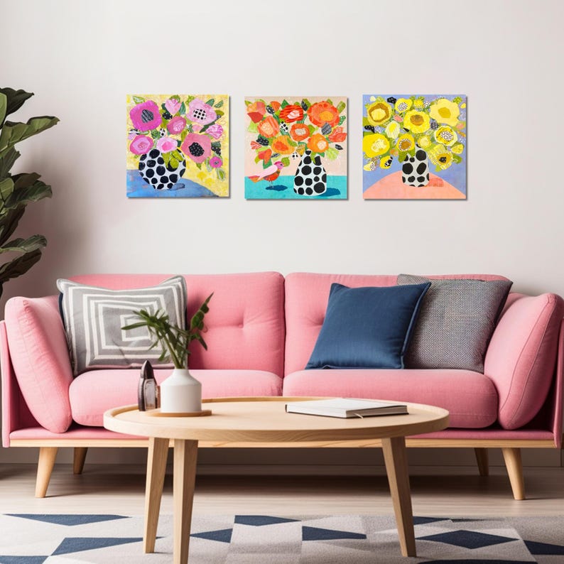 May include: Three colorful floral art prints. Each print features a bouquet of flowers in a black and white polka dot vase. The background colors are yellow, blue, and pink. The prints are displayed above a pink sofa.