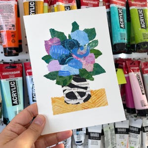May include: A collage art piece featuring a bouquet of flowers in a black and white striped vase. The flowers are made of paper and are in shades of blue, pink, and green. The vase is sitting on a yellow and orange background with text from a book.